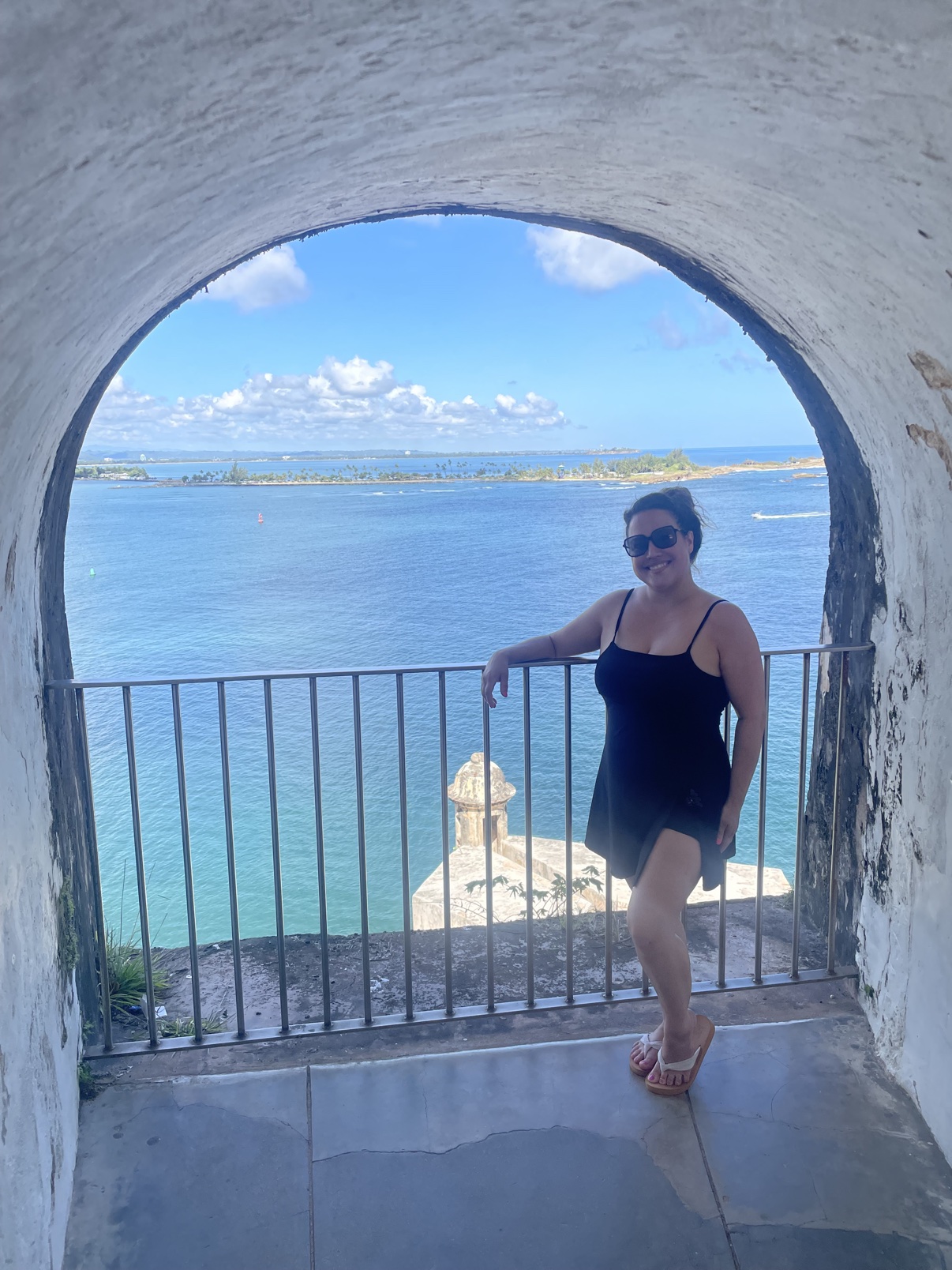 Fatima in a black dress at a stone archway overlooking the ocean